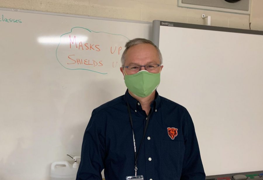 Tony Engle poses in his classroom in front of his masks up, shields up reminder. I had been hesitant to come back because  of Covid-19, being as old as I am, but Im thrilled to be here, Engle said. 